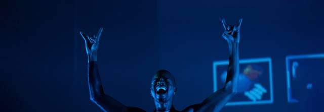 closeup of black male raises his arms in a rocker gesture with his hands with his mouth open in exclamation and his eyes closed. blueish lighting.