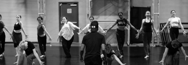 black and white image of dancers in a horizontal line about to take a step forward while raising their arms. a figure faces away from the camera watching.