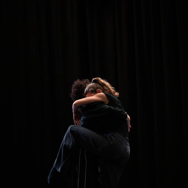 two women wearing dark colored clothing embrace with one dance legs wrapped around the other's body