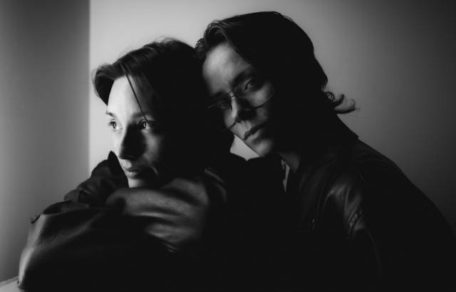 black and white closeup image of two people in a close embrace. one looks at the camera and the other looks away from the camera