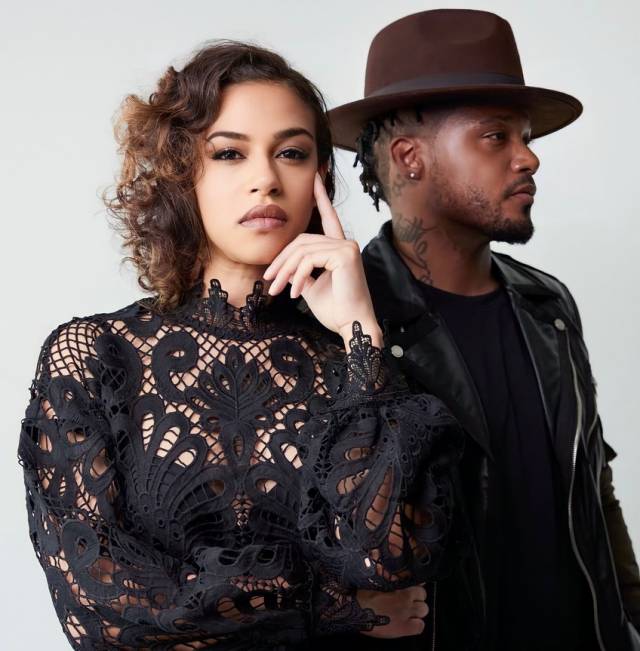 a brown-skinned woman with loose brown curls wearing a black lace top contemplatively looking at the camera and a brown-skinned man with a mustache and beard wearing a black shirt, a black motorcycle jacket, and brown fedora in profile looking off to the side