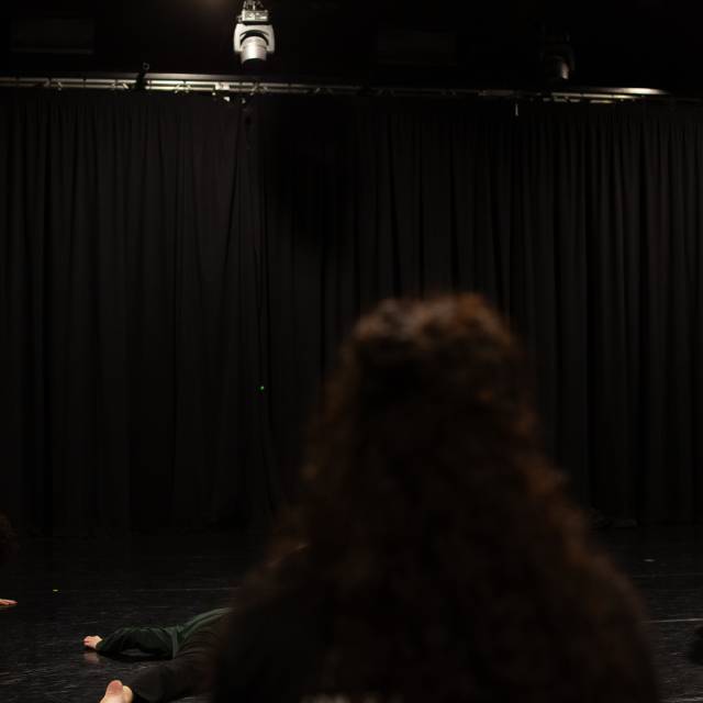 a woman wearing a blue sweater stands on the back of a woman with a grey shirt on all fours. on the opposite side of the image a woman wearing a yellow shirt is captured in motion