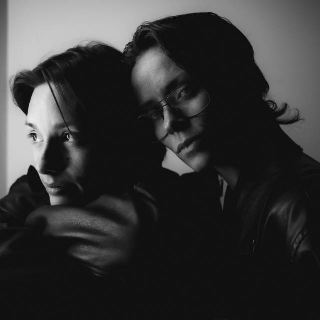 black and white closeup image of two people in a close embrace. one looks at the camera and the other looks away from the camera