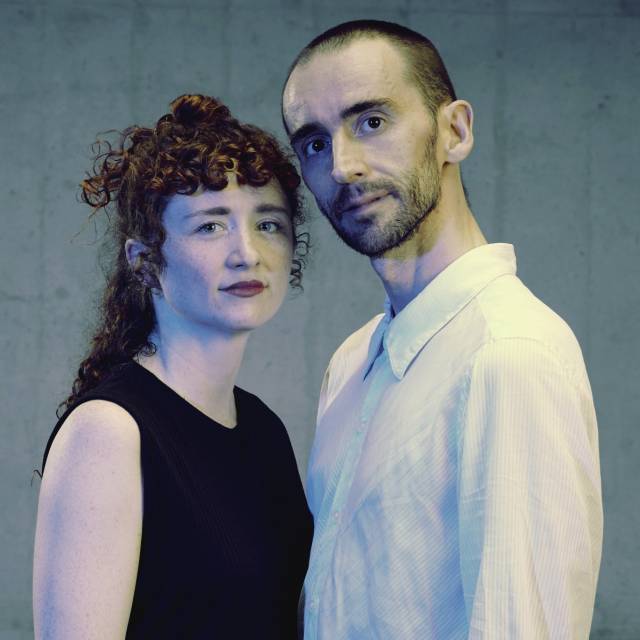 two people look towards the camera. one olive-skinned person has curly red hair and wears a sleeveless black top. the other is olive-skinned with a black mustache and beard and wears a white button down shirt
