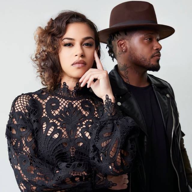 a brown-skinned woman with loose brown curls wearing a black lace top contemplatively looking at the camera and a brown-skinned man with a mustache and beard wearing a black shirt, a black motorcycle jacket, and brown fedora in profile looking off to the side