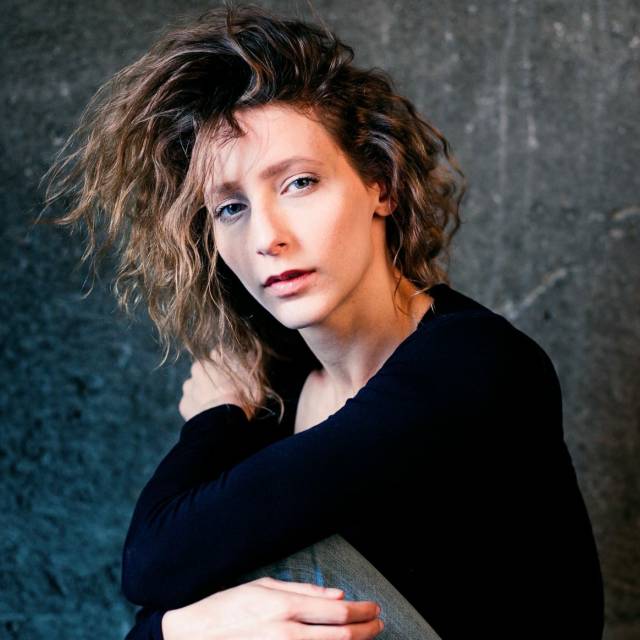 Woman with wavy brownish hair and wearing a black long sleeve shirt looks into camera with her arms loosely wrapped around her