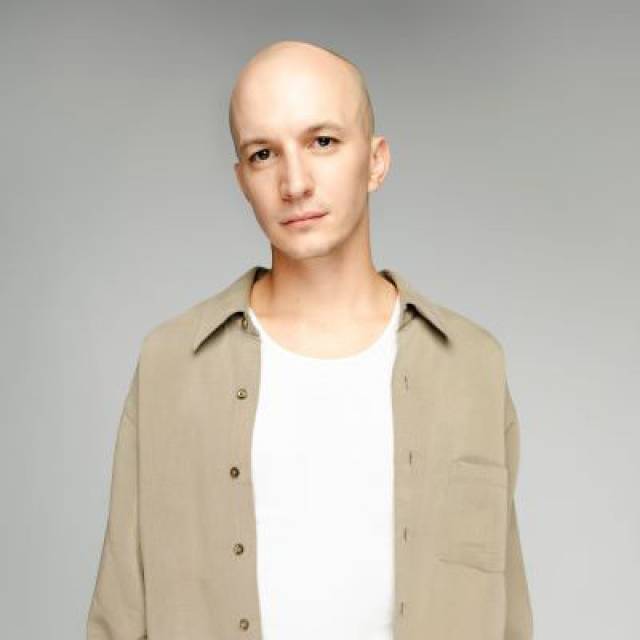 olive skinned man with bald head wearing a white scoop necked shirt and an open long sleeved khaki shirt